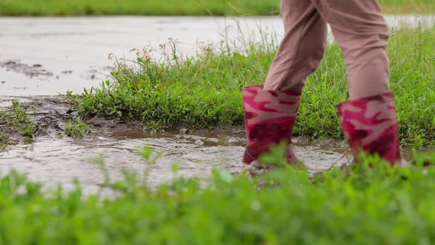 Close-up of a child’s legs in pink floral-patterned rubber boots splashing through a muddy puddle. Water dramatically splashes upward, capturing the joyful energy of play on a rainy day.