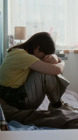 Vertical shot of young woman crying sitting on bed in dorm room with friend entering and supporting her, copy space