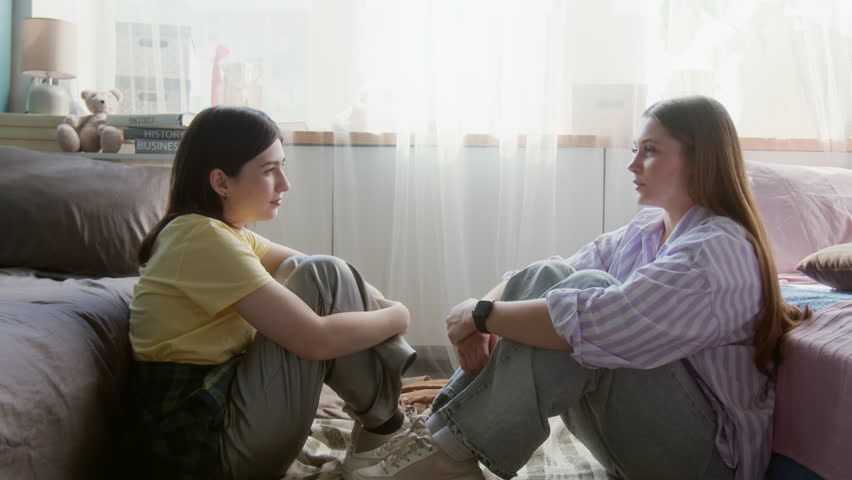 Side view full length shot of two teenage girls sitting opposite each other on floor and chatting sharing intimate stories