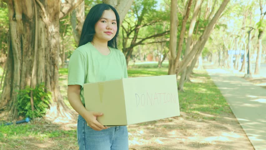 ]Female volunteers hold cardboard boxes in a community park to collect donations to help victims of natural disasters, wars, and the homeless. They enjoy volunteering with a cheerful look.