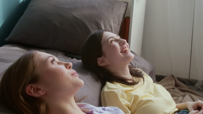 Side view shot of two young teenage girls daydreaming looking up together and chatting while sitting on floor together and laughing