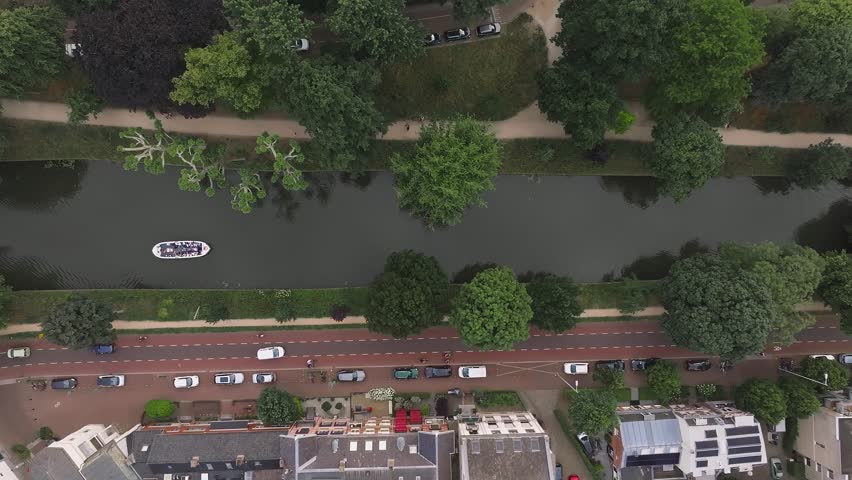 Aerial view of a canal in Utrecht, Netherlands, with a moving boat, green trees, a walking path, parked cars, and solar paneled rooftops.