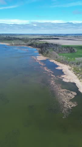 Picturesque vertical aerial of a sprawling lake, shoreline forest, and distant fields. Designed for mobile viewing, social content. Bright, partly cloudy sky illuminates this tranquil rural expanse.