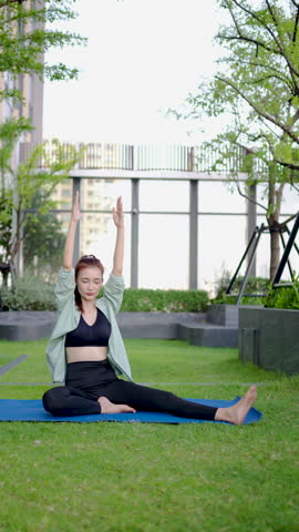 Engaging in a calming yoga session, Adult asian woman sitting on mat stretches her muscles and raises her hands towards the sky. Surrounded by greenery, she enjoys the serenity of her routine