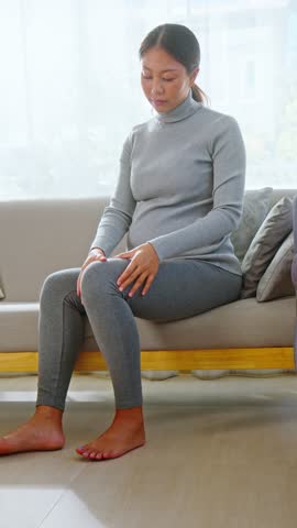 Asian Pregnant woman experiencing knee pain and joint discomfort sitting on sofa at home, health issue and injury concept, female holding knee with pain indoors