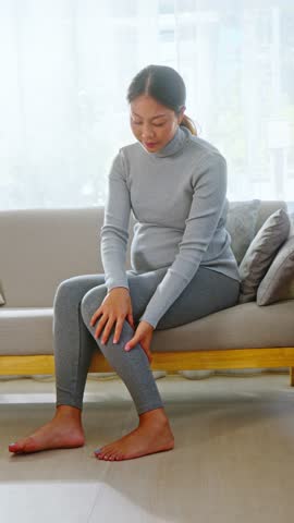 Asian Young pregnant woman experiencing leg pain and discomfort sitting on sofa at home, maternity health issue causing ache and discomfort for pregnant woman