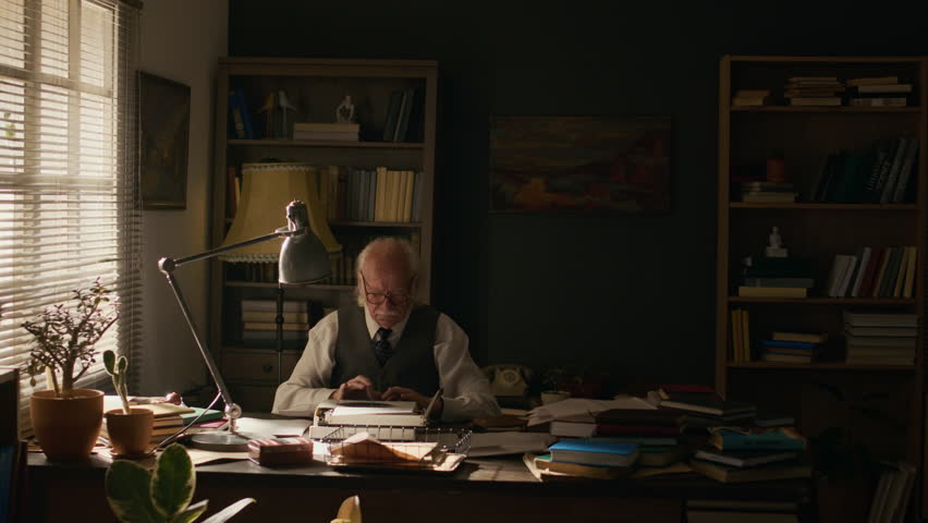 Timelapse shot of elderly Caucasian male writer sitting at desk in his retro home study among pile of books and working on typewriter throughout day