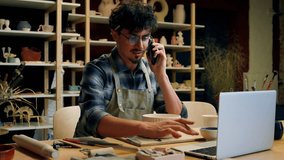 Caucasian man guy potter salesman consult sell ceramics online store small business owner talk mobile phone conversation male sculptor artist typing laptop in pottery workshop craft handmade commerce - Powered by Shutterstock - Get 15% off with code: PIKWIZARD15