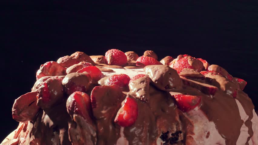 Homemade cake with pink cream decorated with strawberries and liquid chocolate rotates and spreads. Side view on a black background. Festive greeting background