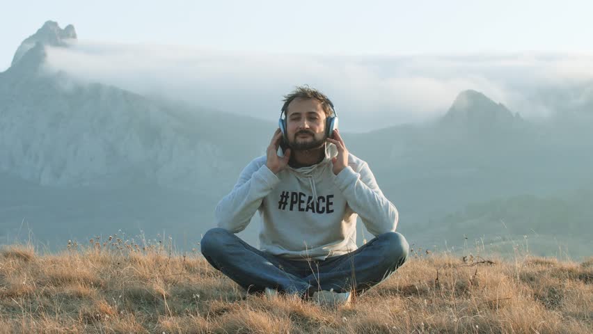 An adult man in jeans and a hat with headphones sits against the backdrop of mountain peaks in the fog and meditates. Vacation mode concept