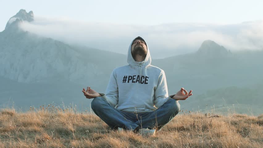 An adult man in jeans and a hat sits against the backdrop of mountain peaks in the fog and meditates. Vacation mode concept