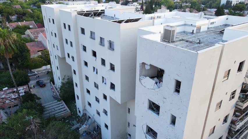 Descending drone shot along a white residential building in Tel Aviv, visibly impacted by a recent strike. A large hole is seen in the structure, with shattered windows, debris on the ground