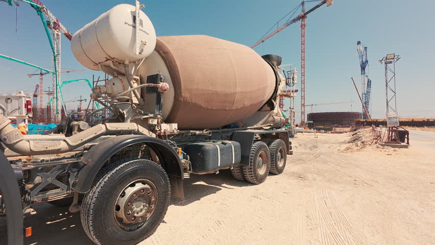 Handheld real-time footage of concrete mixer truck at construction site. Tower cranes at background. Concrete work in progress. Infrastructure, construction, urban development, industrial machinery