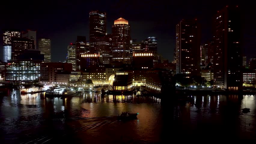 Aerial pullback of Boston Harbor at night, revealing the glowing skyline, waterfront reflections, and the Seaport District in a cinematic urban coastal scene.