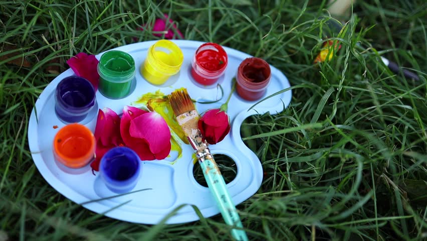 Bright paint containers and a brush sit on a palette surrounded by vibrant petals on the grass, showcasing an outdoor art session filled with creativity and natural beauty.