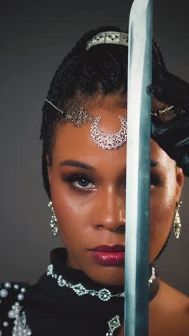 closeup portrait african fantasy woman warrior queen holding sword in hands strokes blade, sexy looks at camera. Beauty face black dress girl princess tiara moon, dark gray studio. vertical video.