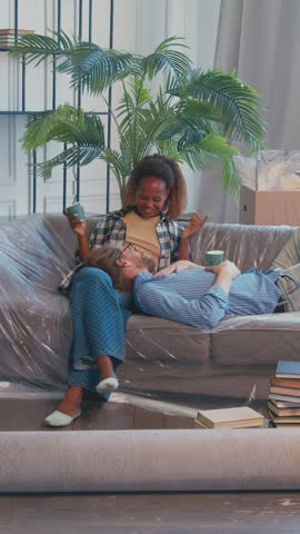 After a long day of moving into a new home, the tired friends take a moment to relax on the couch. Surrounded by boxes and plants, they enjoy some drinks and savor their accomplishment.