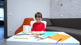 Little toddler kid in red t-shirt sits at desk. Colorful pencils are scattered in front of the child. Baby boy picks a pen and draws on the paper. - Powered by Shutterstock - Get 15% off with code: PIKWIZARD15