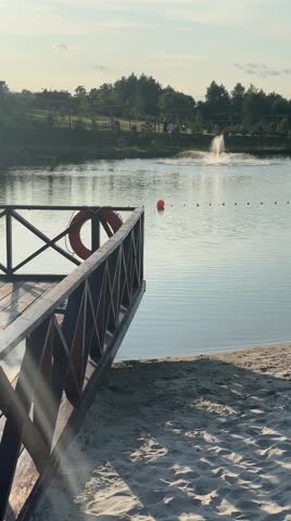 A wooden pier extends over calm lake water at sunset, with a lifebuoy hanging from its railing. A speaker and stage light are visible on the pier, while a fountain sprays water in the distance 
