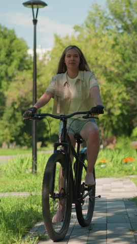 Young woman rides bicycle confidently down paved path in scenic park filled with greenery, sunlight filters through trees, casting dappled shadows on path