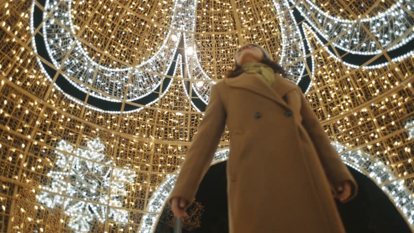 Woman wearing a wool cap and long coat happily looking at a large sphere decorated with Christmas lights.