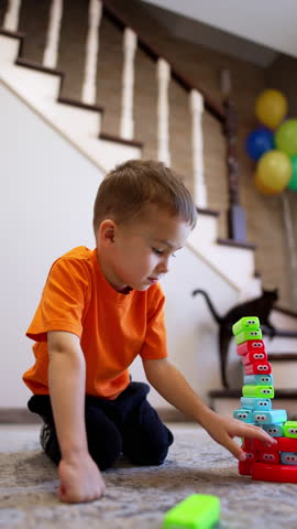 Kid in orange t-shirt puts blocks in a pile and it breaks. Smiling toddler boy sitting on the floor pushes the bricks away. Vertical video.