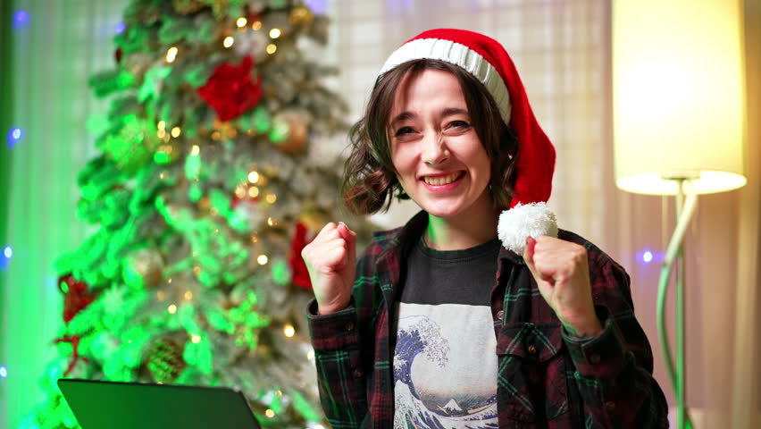 Woman in Santa cap looks at her laptop focused. Girl rejoices clapping hands receiving god news. Christmas tree at backdrop.