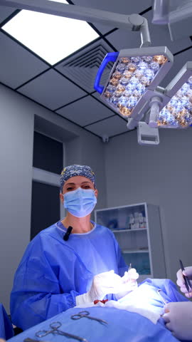 Uniformed female surgeon in flowery cap standing at the operating table talks to the camera. Medics working at operation in modern hospital. Vertical video.