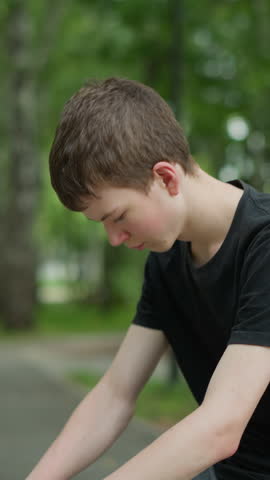 Young boy wearing black top sits with his head down, thoughtfully contemplating on park bench, his bicycle is turned upside down beside him, with a blurry view of trees in the background