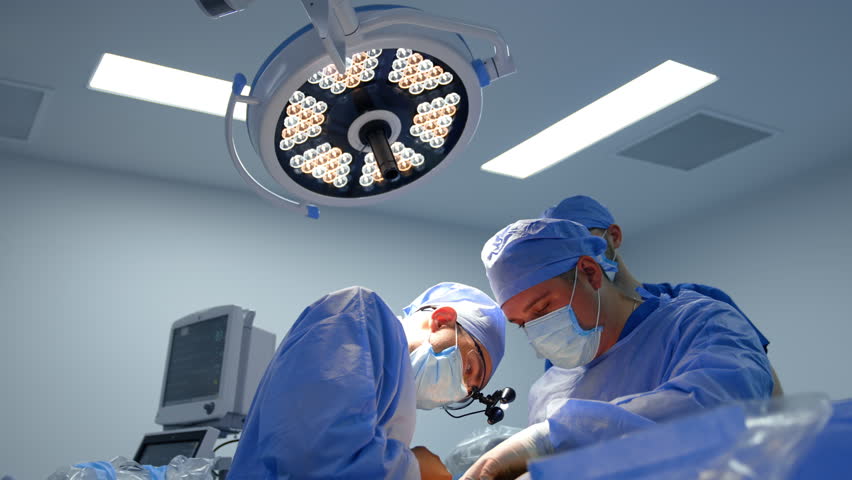 Surgeon in surgical loupes puts efforts to apply a medical instrument. Medical team working in the operating room.