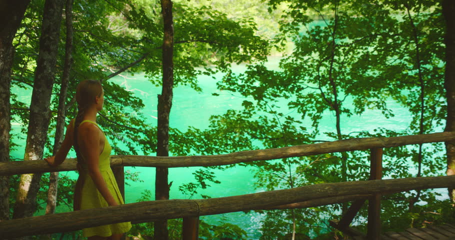 Woman walking on wooden path under shade of trees by emerald lake in Plitvice National Park