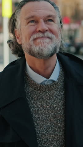 Vertical video, A mature man with a beard and long gray hair smiles as he walks down a city street. He wears a black trench coat and a gray sweater.