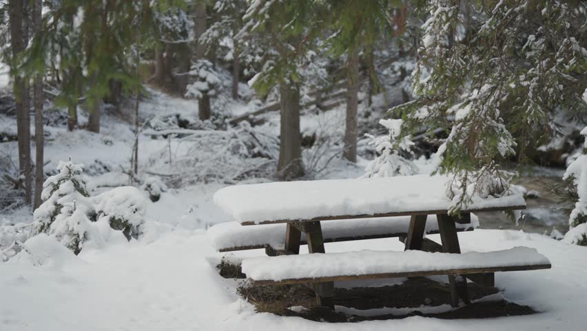 A snow-covered picnic table in the winter forest on the bank of the shallow mountain stream. Parallax video.