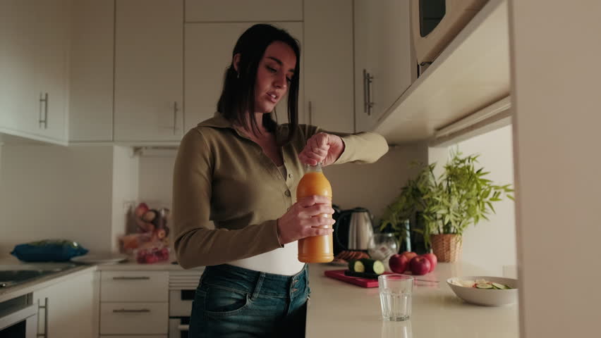A young woman pours herself a glass of orange juice in her modern kitchen. She is wearing casual clothes and has a healthy lifestyle.