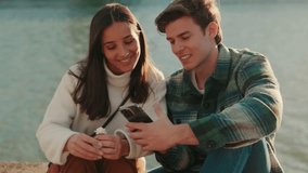 A young couple sits by the river looking at a phone and smiling. The woman holds a small white flower. They are happy and enjoying their time together. - Powered by Shutterstock - Get 15% off with code: PIKWIZARD15