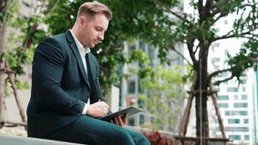 Side view of businessman sitting at park while writing on tablet by using tablet pencil. Low angle camera of smart project manager looking at data analysis while thinking about financial plan. Urbane - Powered by Shutterstock - Get 15% off with code: PIKWIZARD15