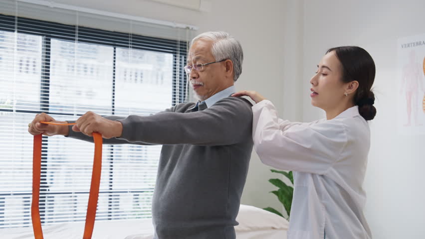  Asian female doctor or physiotherapist guides her elderly male patient through shoulder muscle exercises using a medical resistance band. She carefully monitors posture and offers hands-on support 