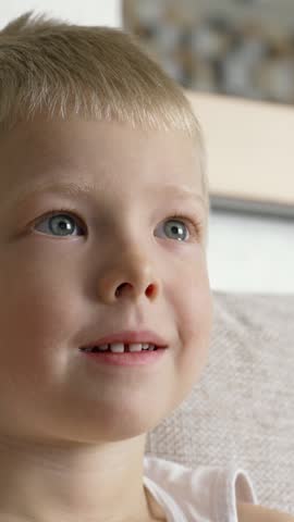 Blond preschooler sitting comfortably on living room sofa, watching television with changing facial expressions, showcasing childhood curiosity and engagement