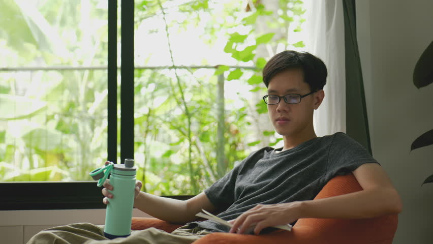 Asian man enjoys reading a book by the window using natural light and drinking from a reusable bottle.4k