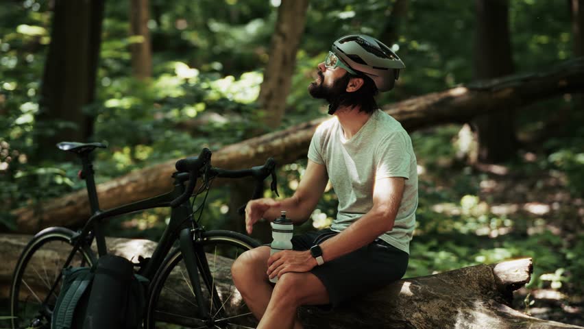 Cyclist drinks water from bottle after intense MTB training. Athletic man hydrates during triathlon cycling. Sporty lifestyle, Cyclist endurance recovery in outdoor mountain biking. Cyclist isotonic drinks. Cyclist Rest. Effort Cycling In Forest