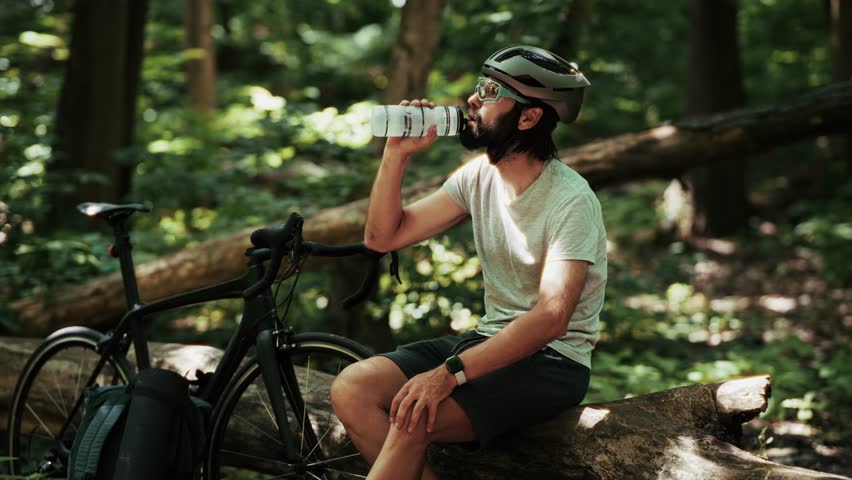 Cyclist drinks water from bottle after intense MTB training. Athletic man hydrates during triathlon cycling. Sporty lifestyle, endurance recovery in outdoor mountain biking. Cyclist isotonic drinks