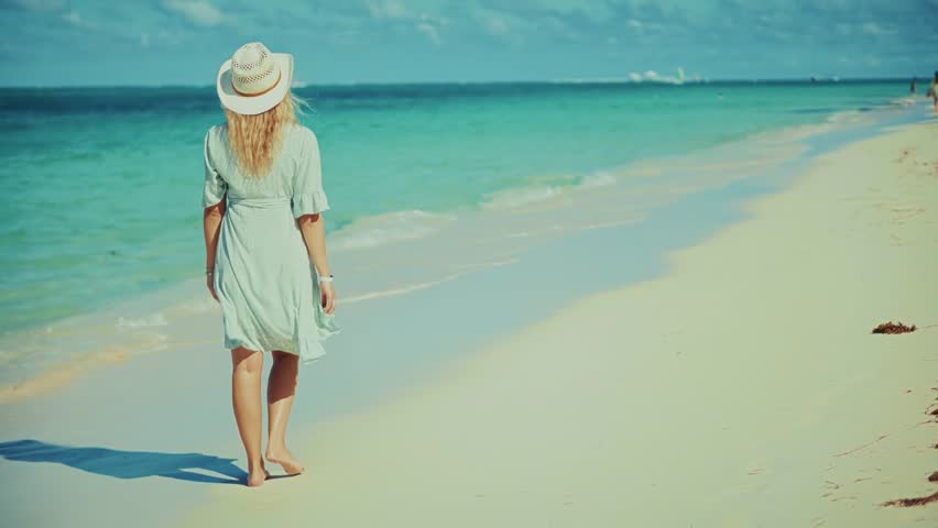 lone woman in a white dress and straw hat walks barefoot along a pristine sandy beach, gazing out at the vast turquoise ocean under a clear sky, embodying tranquility and vacation
