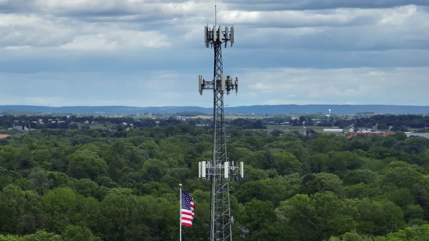 BLowing american flag on flagpole beside transmission tower in rural area with green trees. Aerial close up shot. Cloudy day with Agricultural farm fields in distance.