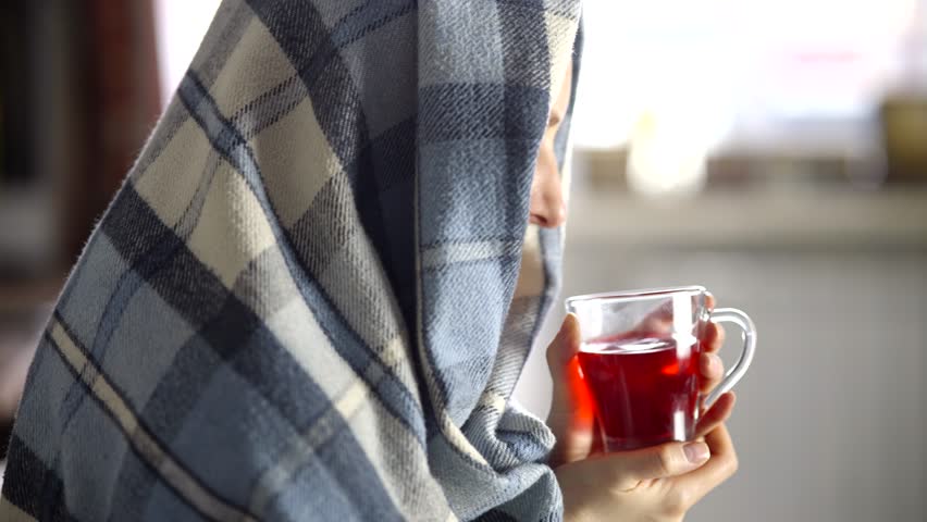 sick woman with sinus congestion drinks hot tea for relief. Wrapped in warmth, she battles seasonal flu and rhinitis. Comfort, medicine, and rest help soothe her severe cold and infection symptoms