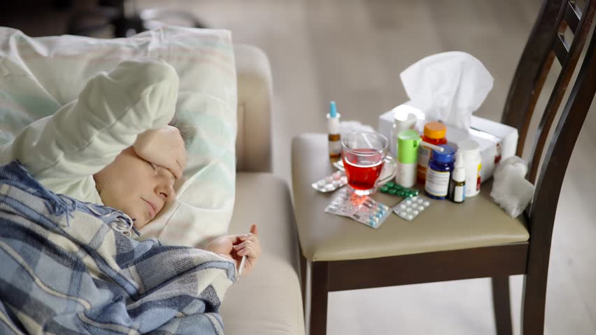 A sick person lies in bed, with a hand on their forehead, while a nearby chair holds collection of medicines, tissues, and a drink, indicating illness. showing discomfort col symptoms headache relief