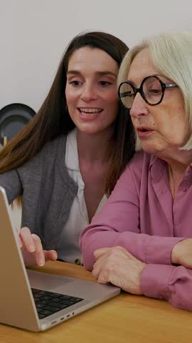Young woman teaching her grandmother how to use laptop computer sitting together in the kitchen at home, enjoying quality time and connection in their home while drinking homemade orange juice 