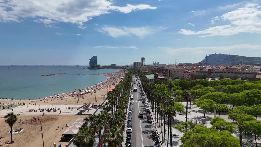 An energetic aerial shot showcases Barceloneta Beach in Barcelona, Spain, bustling with sunbathers and framed by a palm-lined promenade and the city skyline.