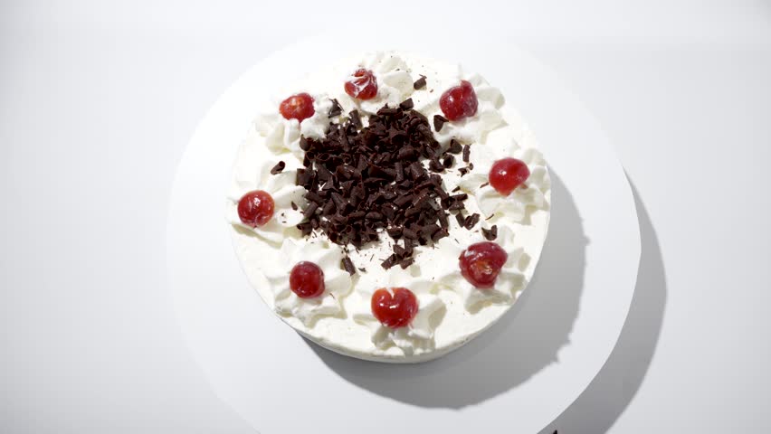Whole cake from above. A round cake with cream, cherries, and chocolate shavings rotates from a bird