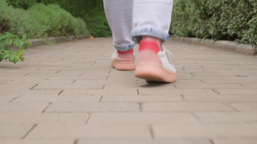Feet in sneakers walking along path between green hedges in park.