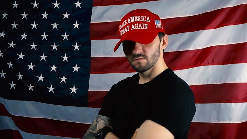 Patriotic white man with tattoos and beard putting on a red MAGA or Make America Great Again hat in slow motion in front of a waving american flag and looks directly at the camera with confidence.